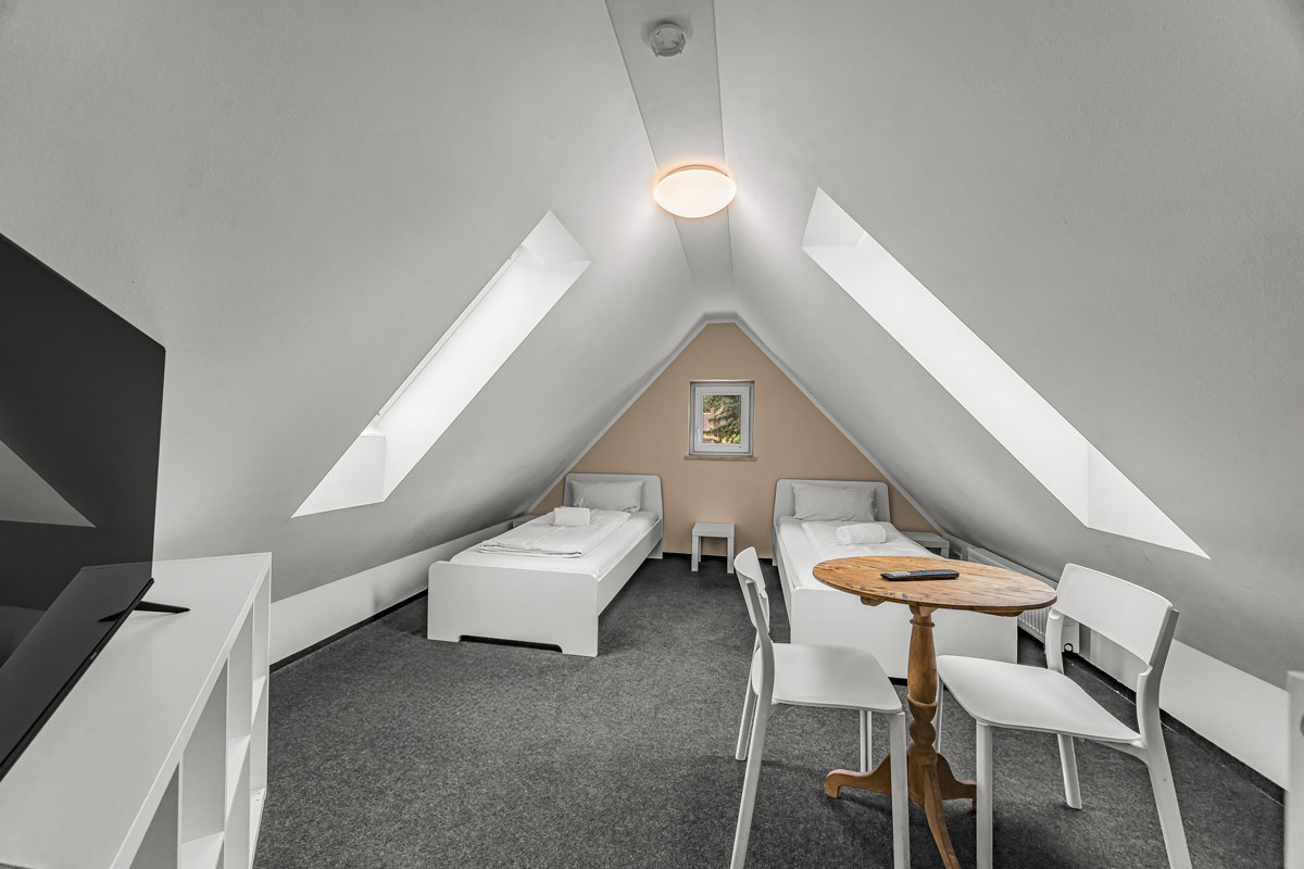 Ein helles Dachzimmer mit zwei weißen Betten und einem runden Holztisch.