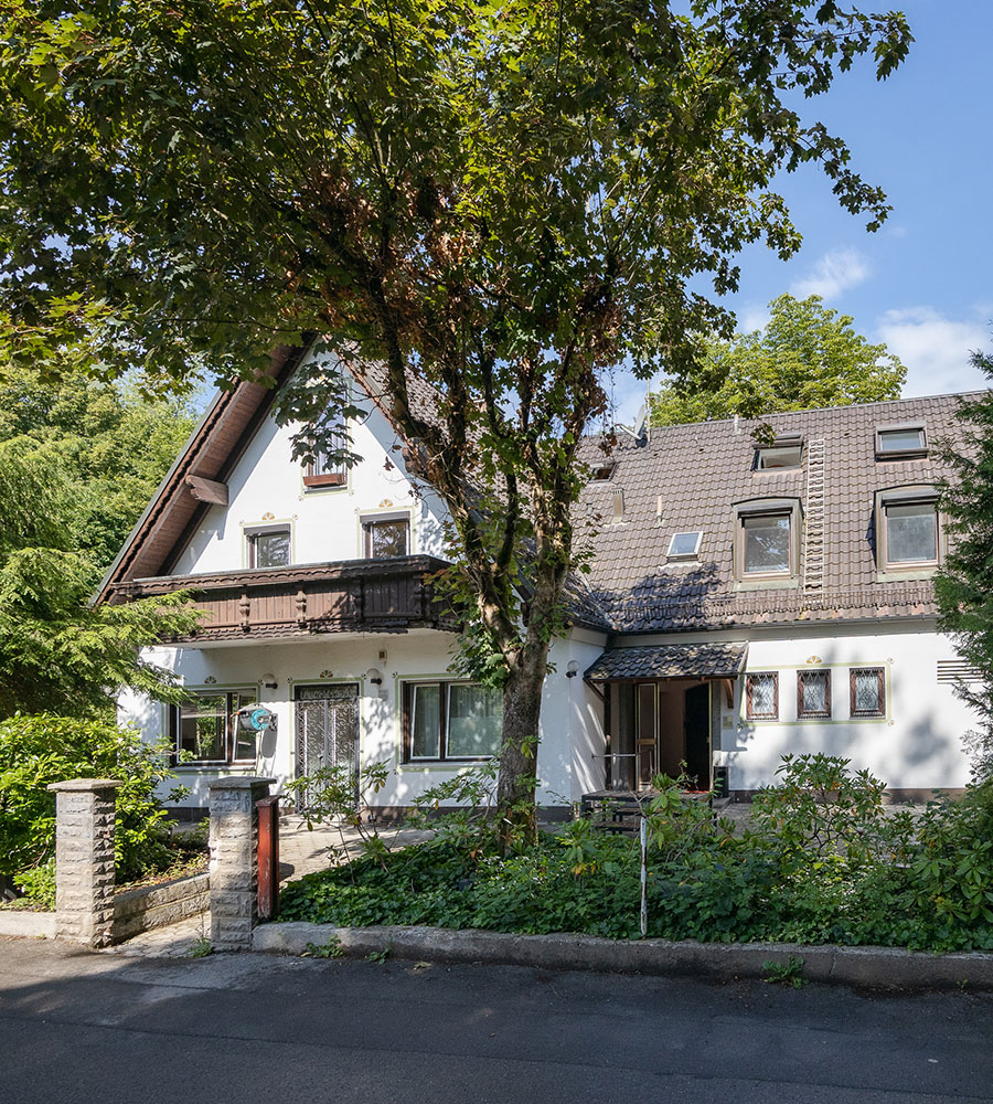 Zweistöckiges Haus mit Schrägdach und großem Baum im Vordergrund.