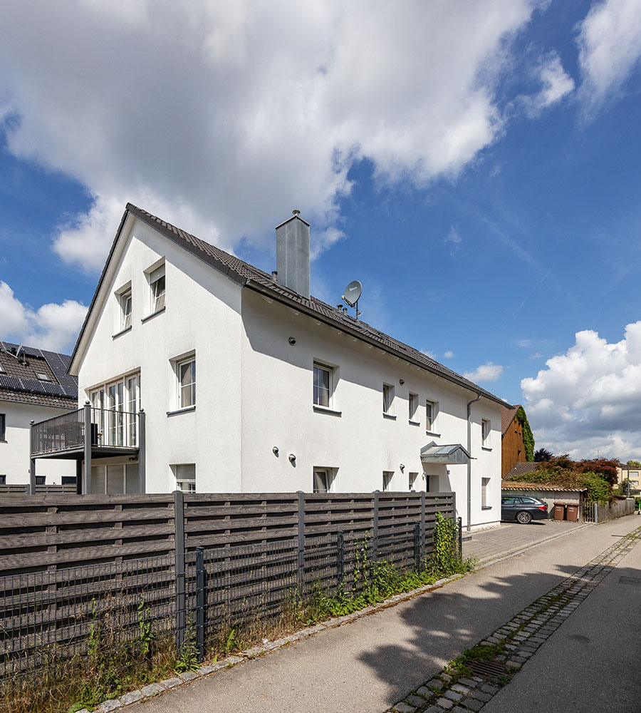Zweistöckiges weißes Haus, blauer Himmel, Zaun vorne, gepflasterter Gehweg.