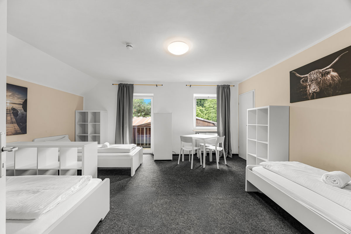 Ein heller Raum mit drei weißen Betten, Tisch und Regalen, Blick nach draußen.