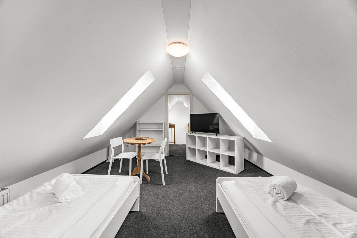 Minimalistischer Dachbodenraum mit zwei Betten, Tisch, Stühlen, helle Farben.
