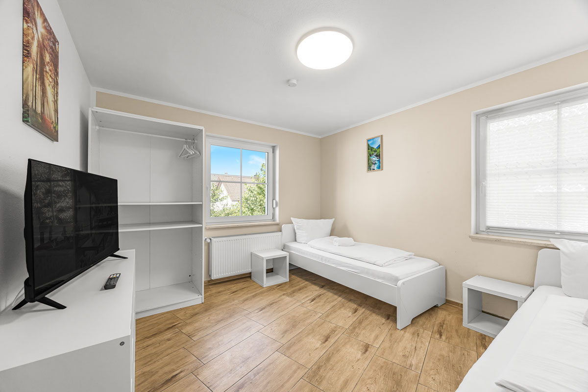 Helles Zimmer mit Holzboden, zwei Betten, TV und offenem Schrank.