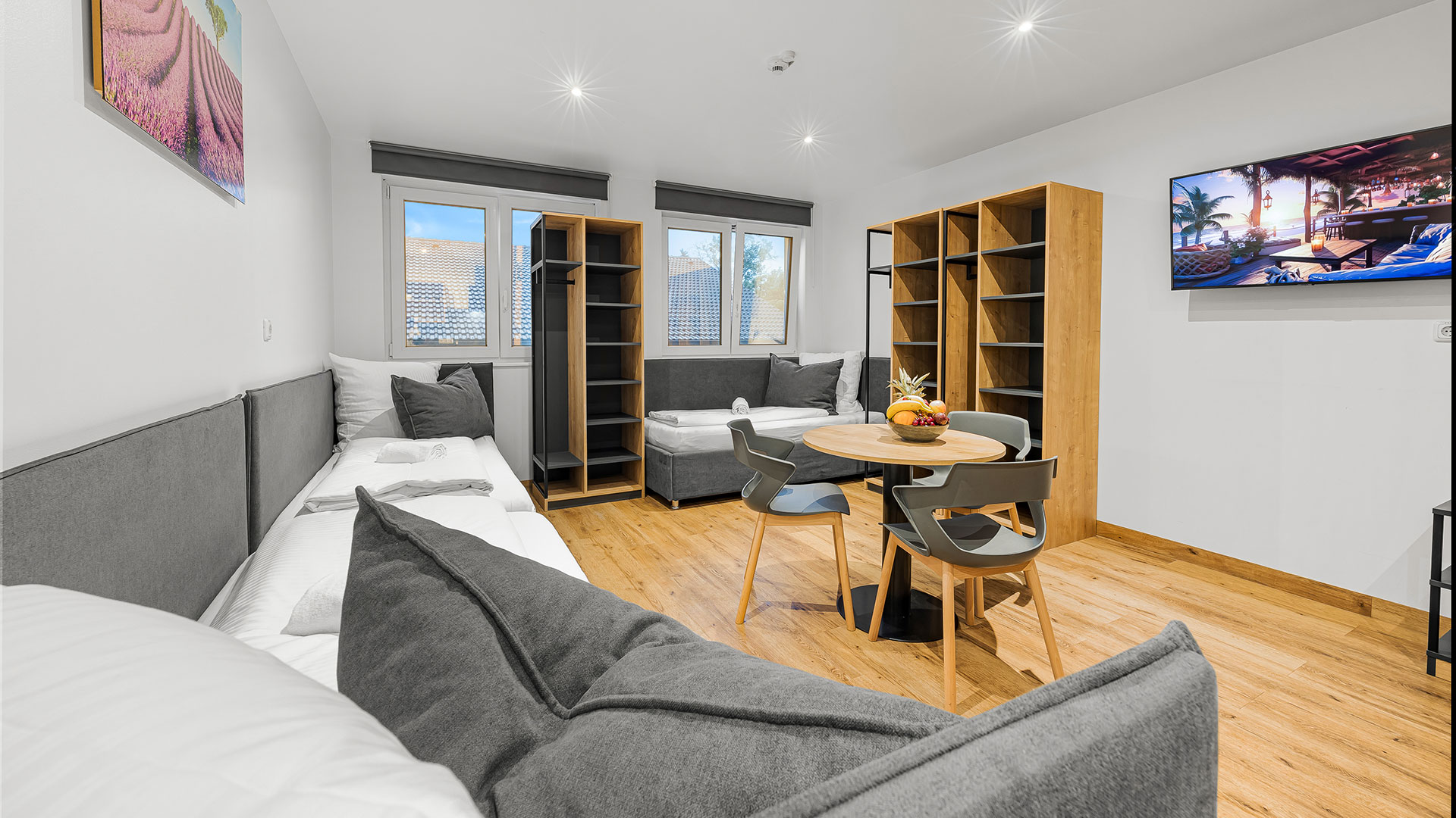 Helles Wohnzimmer mit grauen Sofas, Holzboden, Tisch, Stühlen und Fernseher.