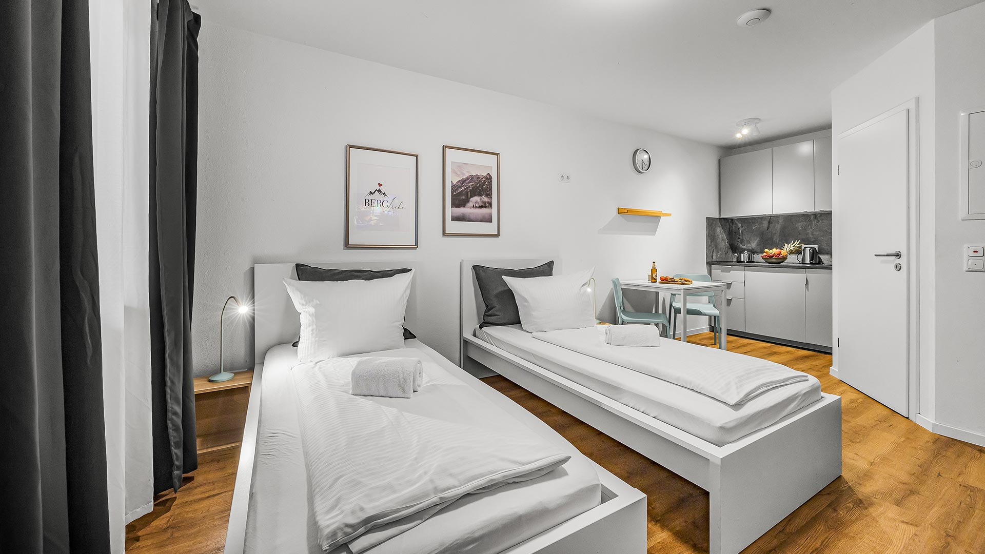 Helles, modernes Zimmer mit zwei Einzelbetten, Küche und Holzboden, dekorativ eingerichtet.