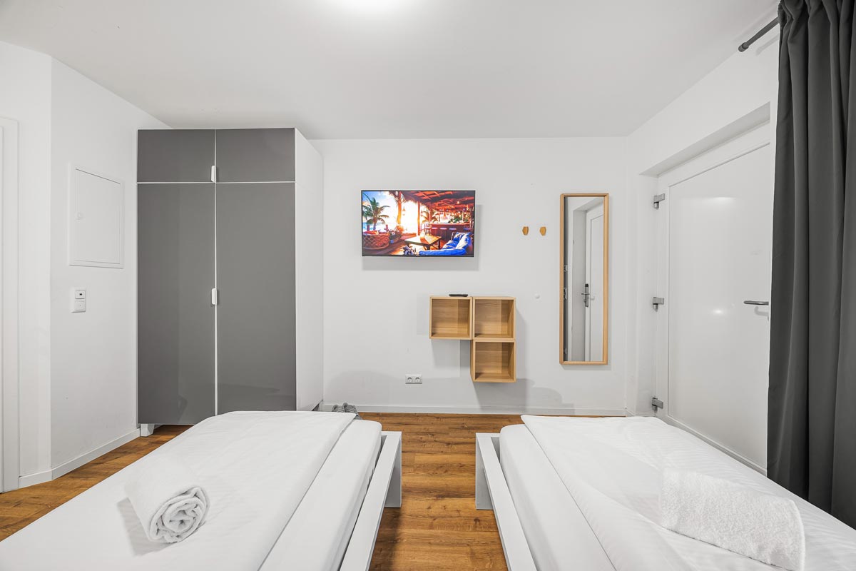 Zwei Betten mit weißen Bettlaken, grauer Schrank und hölzerner Bodenbelag.