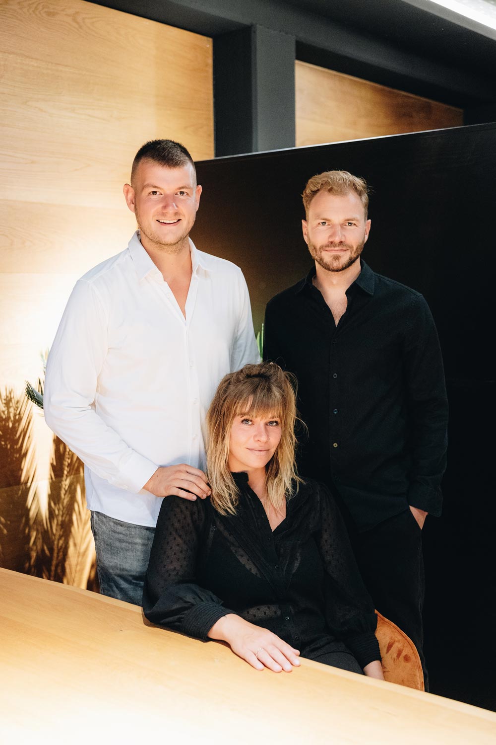 Drei Personen, ein Mann in weißem Hemd, ein Mann und eine Frau in schwarz, lächelnd.